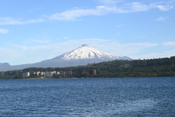 Sicht auf den aktiven Vulkan bei Villarrica mit See im Vordergrund bei schönem Wetter, die Bergspitze ist dabei mit Schnee bedeckt