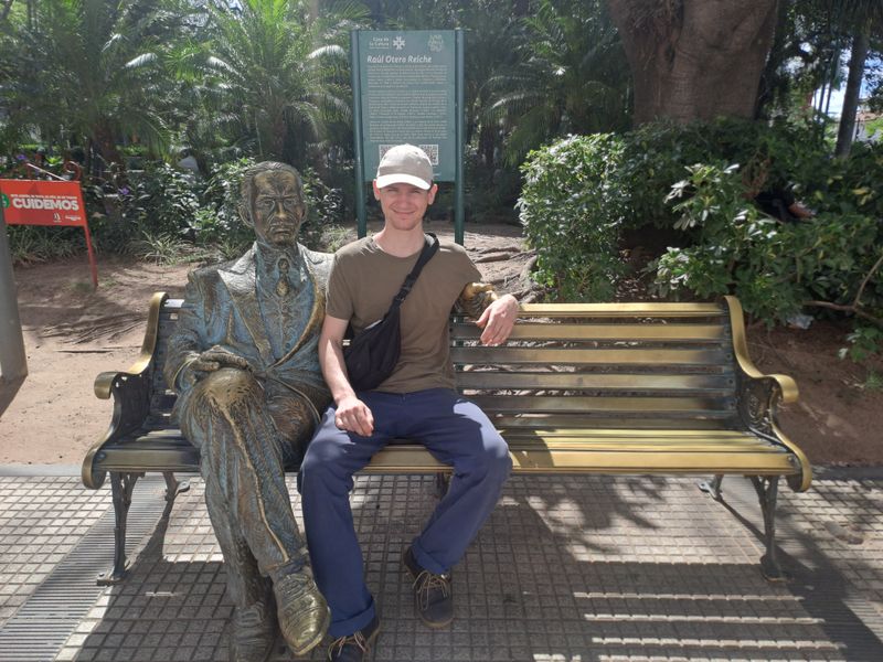 Pascal sitzt auf einer Bank neben einer lebensgroßen Bronzefigur.